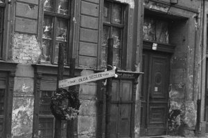 Auf dem Gehweg vor einer heruntergekommenen Häuserfassade mit zugemauerten Fenstern steht ein Mahnmal. Es besteht aus drei mit Stacheldraht umwickelten Pfählen, zwei Kränzen und einem Schild mit Olga Seglers Namen, Geburts- und Todesdatum.