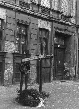 Auf dem Gehweg vor einer heruntergekommenen Häuserfassade mit zugemauerten Fenstern steht ein Mahnmal. Es besteht aus drei mit Stacheldraht umwickelten Pfählen, zwei Kränzen und einem Schild mit Olga Seglers Namen, Geburts- und Todesdatum.