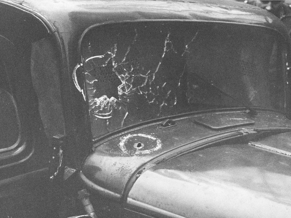 Aufnahme der Windschutzscheibe, die auf der linken Seite von einem Einschuss zersplittert ist. In der Motorhaube ist ein weiteres Einschussloch.