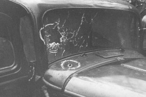 Aufnahme der Windschutzscheibe, die auf der linken Seite von einem Einschuss zersplittert ist. In der Motorhaube ist ein weiteres Einschussloch.
