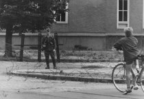 Frontal fotografiert ist ein bewaffneter Soldat hinter einer Stacheldraht-Absperrung. Im rechten Bildvordergrund steht ein Junge mit Fahrrad mit dem Rücken zur Kamera und blickt in Richtung des Soldaten. Im Bildhintergrund ist eine Häuserfassade zu sehen.