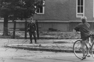 Frontal fotografiert ist ein bewaffneter Soldat hinter einer Stacheldraht-Absperrung. Im rechten Bildvordergrund steht ein Junge mit Fahrrad mit dem Rücken zur Kamera und blickt in Richtung des Soldaten. Im Bildhintergrund ist eine Häuserfassade zu sehen.