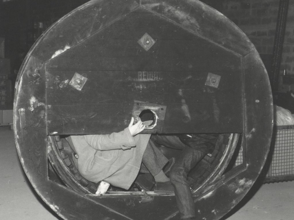 Eine Kabelrolle ist zentral im Bild. Ein Teil der Abdeckung fehlt und man erkennt die untere Hälfte von zwei Männern, die im Inneren der Rolle sitzen. Einer hält den oberen Teil der Abdeckung fest, der ihre Oberkörper verdeckt.
