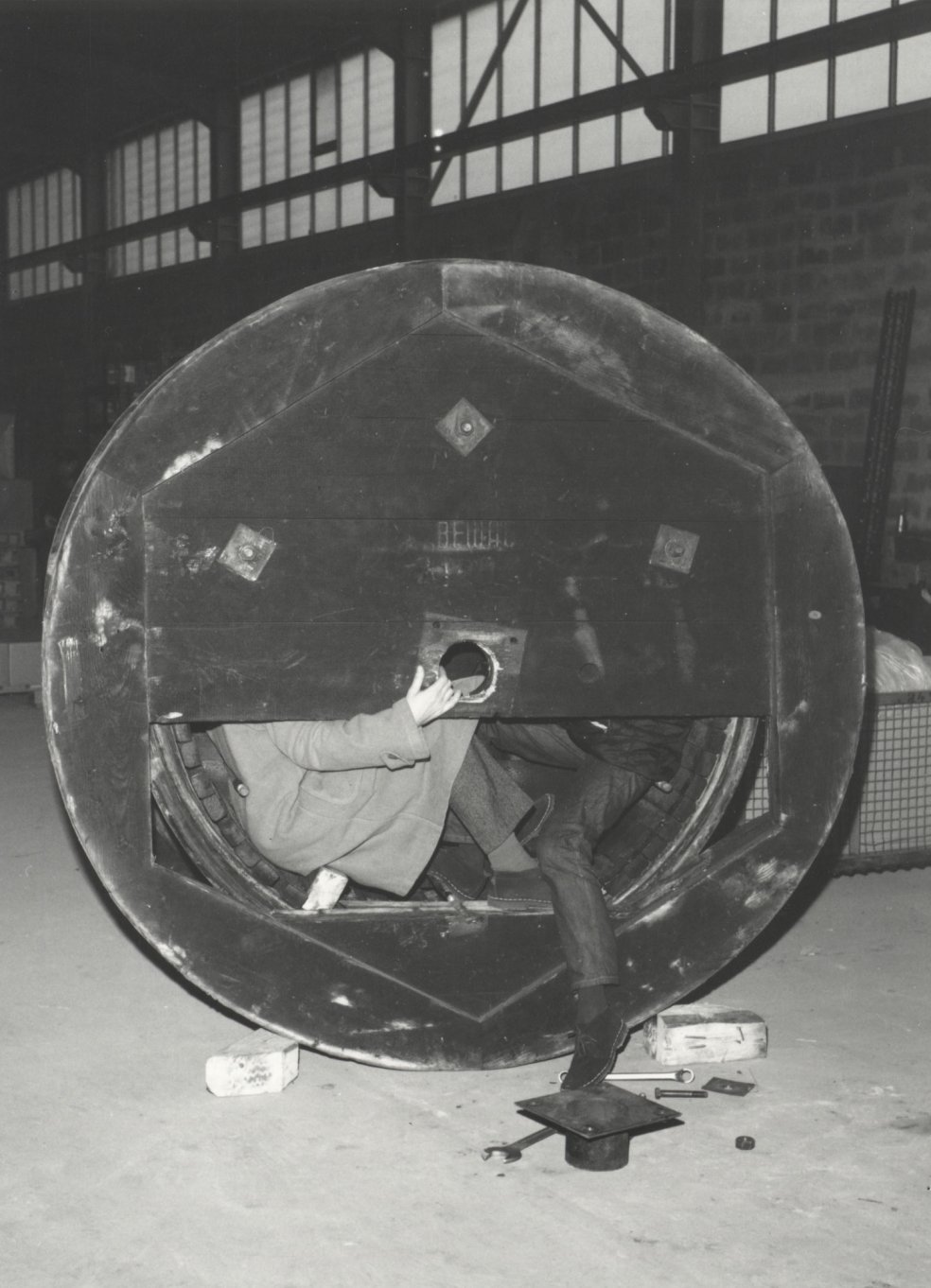 Eine Kabelrolle ist zentral im Bild. Ein Teil der Abdeckung fehlt und man erkennt die untere H&auml;lfte von zwei M&auml;nnern, die im Inneren der Rolle sitzen. Einer h&auml;lt den oberen Teil der Abdeckung fest, der ihre Oberk&ouml;rper verdeckt.