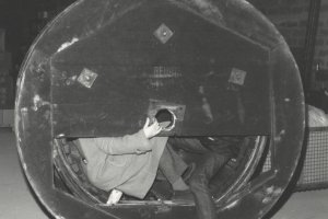 Eine Kabelrolle ist zentral im Bild. Ein Teil der Abdeckung fehlt und man erkennt die untere Hälfte von zwei Männern, die im Inneren der Rolle sitzen. Einer hält den oberen Teil der Abdeckung fest, der ihre Oberkörper verdeckt.