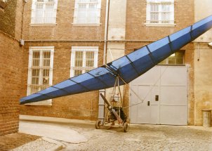 Ein Fluggleiter steht aufgebaut vor einer Gefängniswand. Er hat drei kleine Räder, einen schmalen Propeller und einen Sitz am Gestell, darüber ist der große, blaue Tragflügel ausgebreitet.