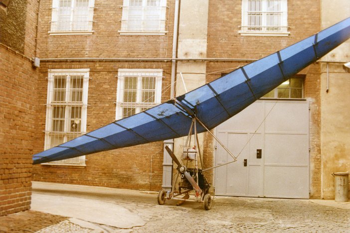 Ein Fluggleiter steht aufgebaut vor einer Gefängniswand. Er hat drei kleine Räder, einen schmalen Propeller und einen Sitz am Gestell, darüber ist der große, blaue Tragflügel ausgebreitet.