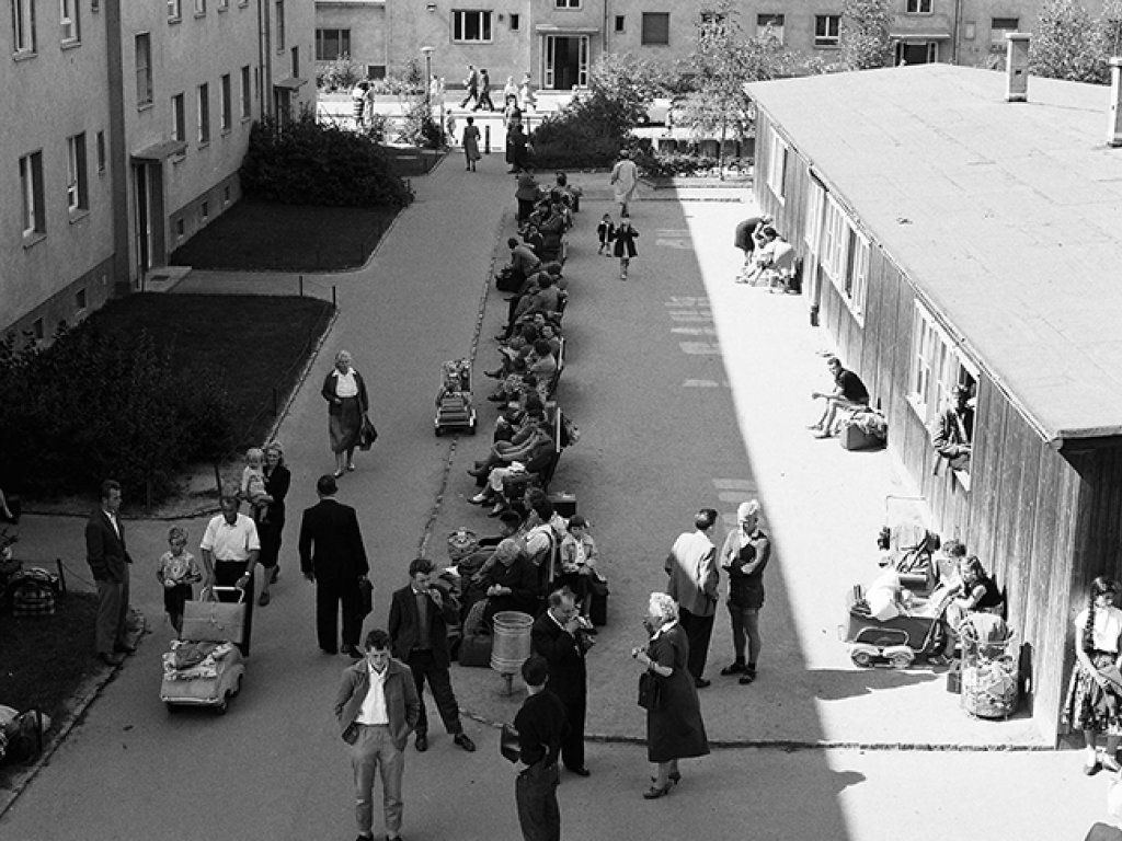Der Blick geht von oben auf eine Straße, die zwischen zwei mehrstöckigen Gebäuden und einer Baracke liegt. In der Mitte stehen Menschen oder sitzen auf Bänken, einige mit Gepäck und Kinderwägen.