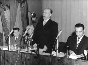 Walter Ulbricht steht in der Bildmitte und spricht in das vor ihm stehende Mikrofon. Rechts und links neben ihm sitzen drei Männer, die in Unterlagen blättern. Im linken Bildhintergrund sind DDR-Fahnen drapiert.