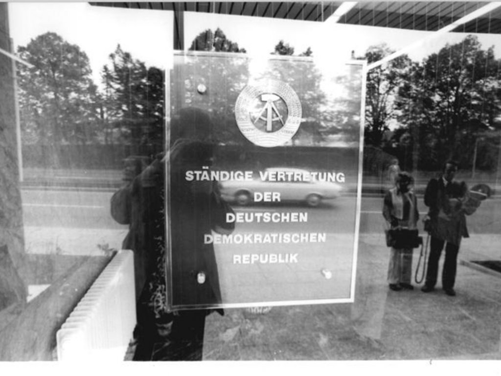 Durch eine Scheibe fotografiertes Schild mit dem Emblem der DDR und der Aufschrift: Ständige Vertretung der Deutschen Demokratischen Republik.
