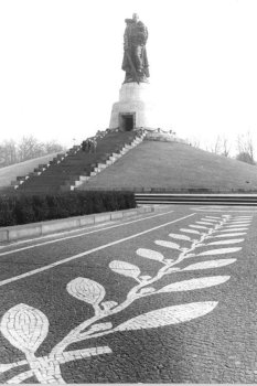 Das Ehrenmal ist von unten fotografiert, die Statue des Soldaten auf einem Sockel am Ende einer Treppe ist im oberen Bildviertel. Groß im Vordergrund ist die weiße Zierpflasterung am Boden.