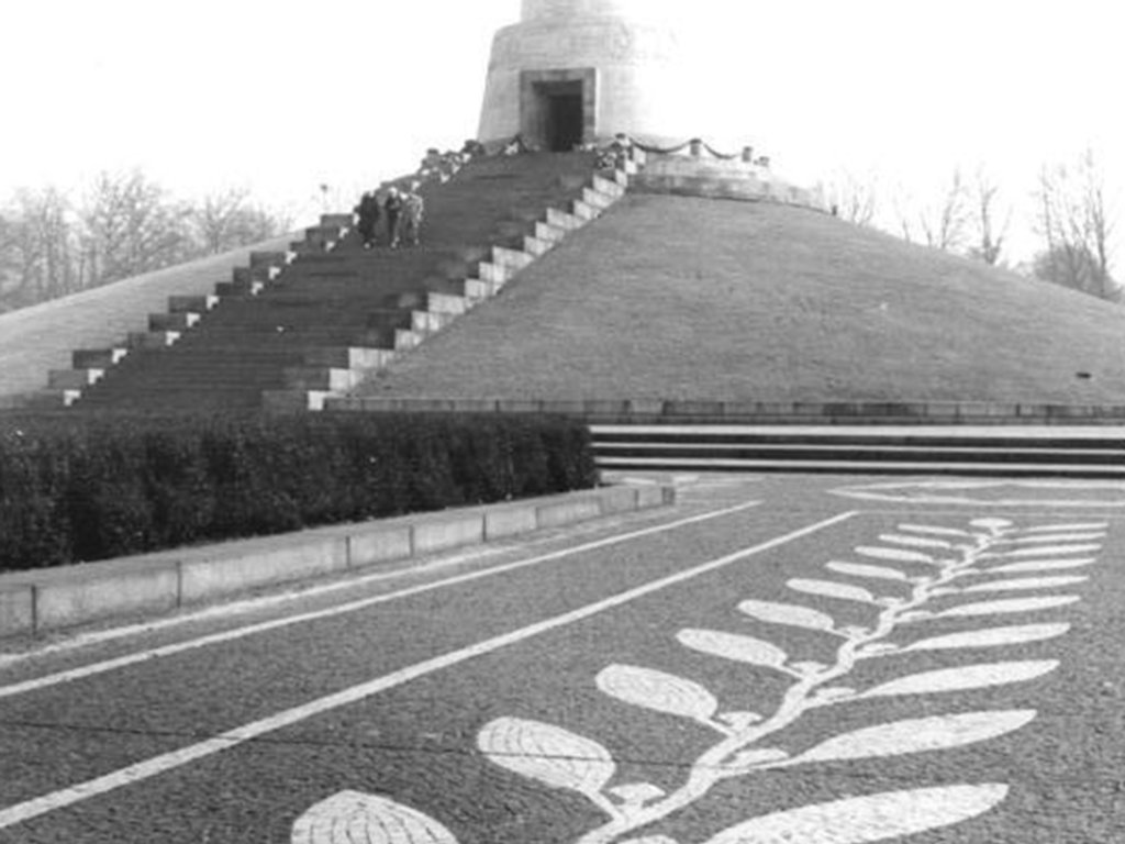 Das Ehrenmal ist von unten fotografiert, die Statue des Soldaten auf einem Sockel am Ende einer Treppe ist im oberen Bildviertel. Groß im Vordergrund ist die weiße Zierpflasterung am Boden.
