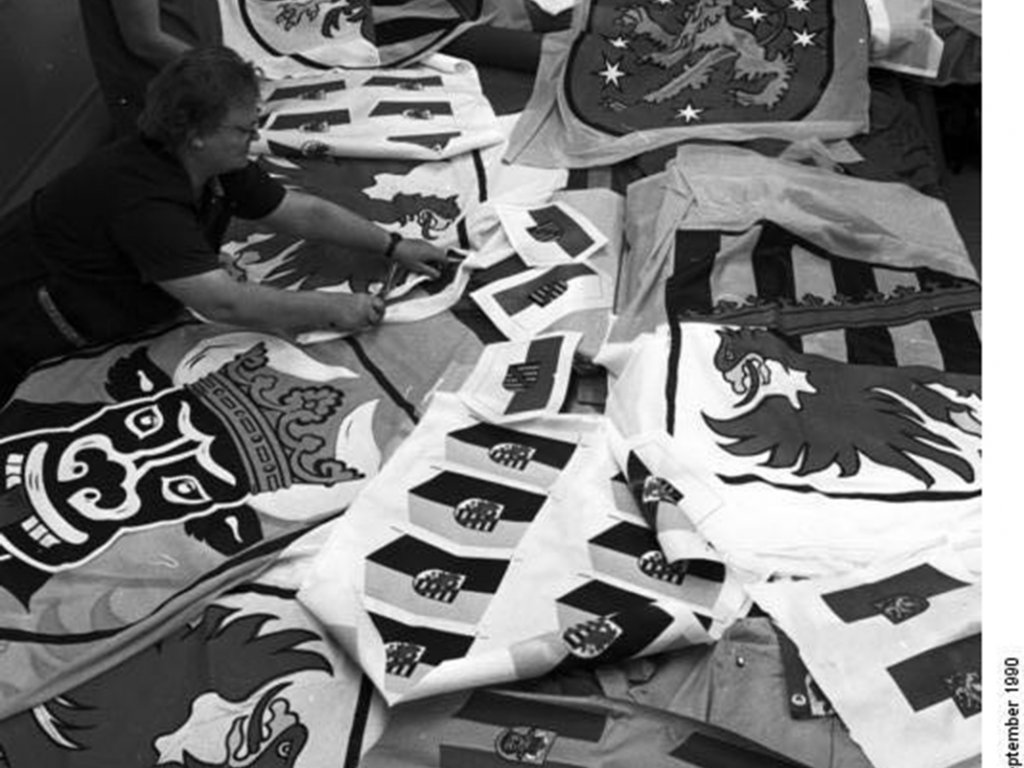 Herstellung der neuen Länderflaggen für die neuen Bundesländer in der Spectrum-Näherei in Leipzig; Aufnahme 20. September 1990  Im Vordergrund liegen zahlreiche Flaggen auf einem Tisch, unter anderem von Brandenburg, Mecklenburg-Vorpommern, Sachsen und Thüringen. Am Tischende arbeiten vier Näherinnen.