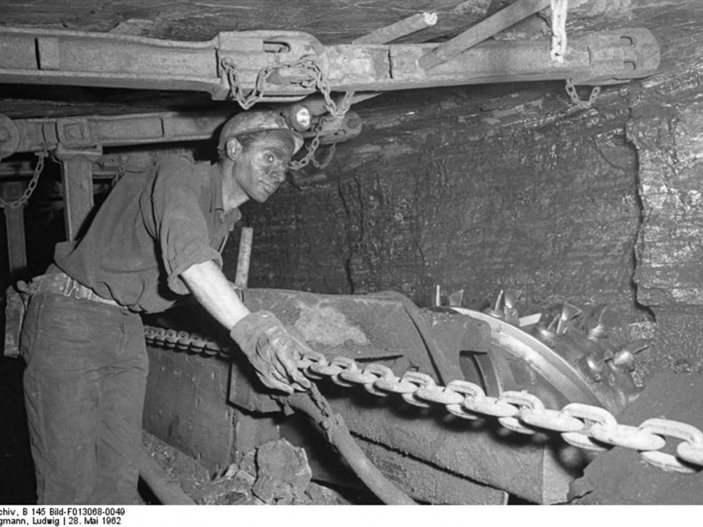 Ein Bergmann unter Tage mit Helm, Handschuhen und verrußtem Gesicht zieht an einer Kette. Die niedrige Stollendecke über ihm ist mit Eisenträgern abgestützt.
