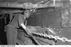 Ein Bergmann unter Tage mit Helm, Handschuhen und verrußtem Gesicht zieht an einer Kette. Die niedrige Stollendecke über ihm ist mit Eisenträgern abgestützt.