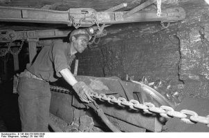 Ein Bergmann unter Tage mit Helm, Handschuhen und verrußtem Gesicht zieht an einer Kette. Die niedrige Stollendecke über ihm ist mit Eisenträgern abgestützt.