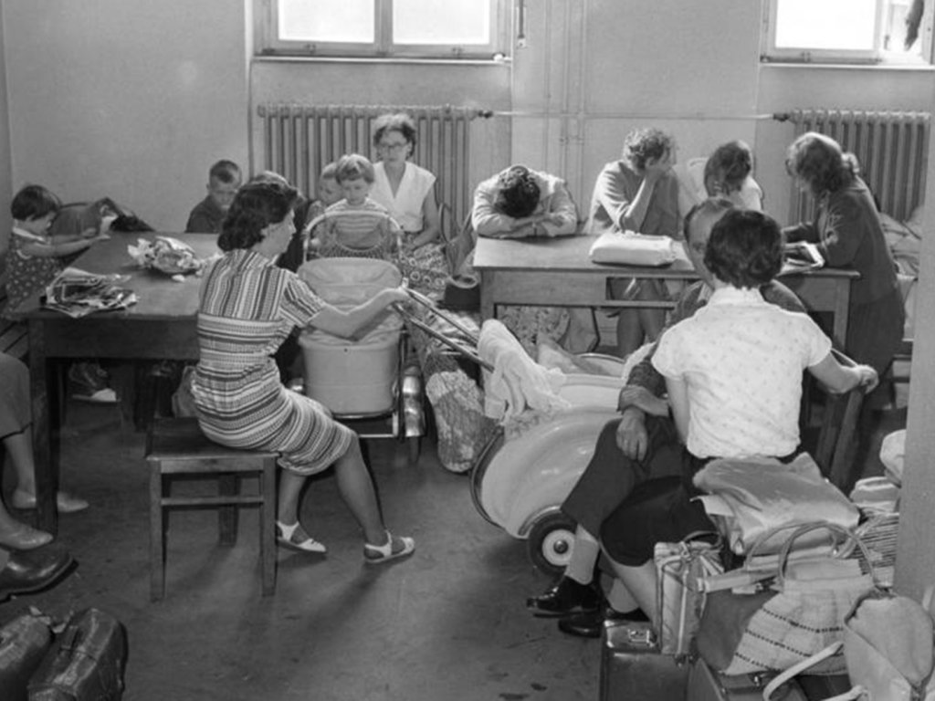 DDR-Flüchtlinge im Notaufnahmelager Marienfelde in West-Berlin; Aufnahme 13. Juli 1961 Ein Raum voller Menschen. Sie sitzen auf Bänken und Hockern, mehrere Kinder und Kinderwagen sind darunter. An einem Tisch sitzen Frauen, die ihre Köpfe in die Hände gestützt oder auf ihre Arme gelegt haben. Im Vordergrund stehen Gepäckstücke.