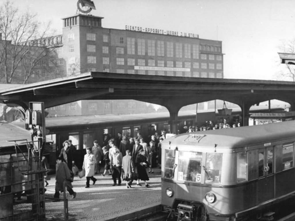Man blickt auf zwei Züge und einen Bahnsteig voller Menschen. Auf dem Dach des Gebäudes im Hintergrund ist eine Flagge zu erkennen und der Schriftzug Elektroapperatewerke J. W. Stalin