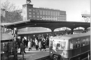 Man blickt auf zwei Züge und einen Bahnsteig voller Menschen. Auf dem Dach des Gebäudes im Hintergrund ist eine Flagge zu erkennen und der Schriftzug Elektroapperatewerke J. W. Stalin