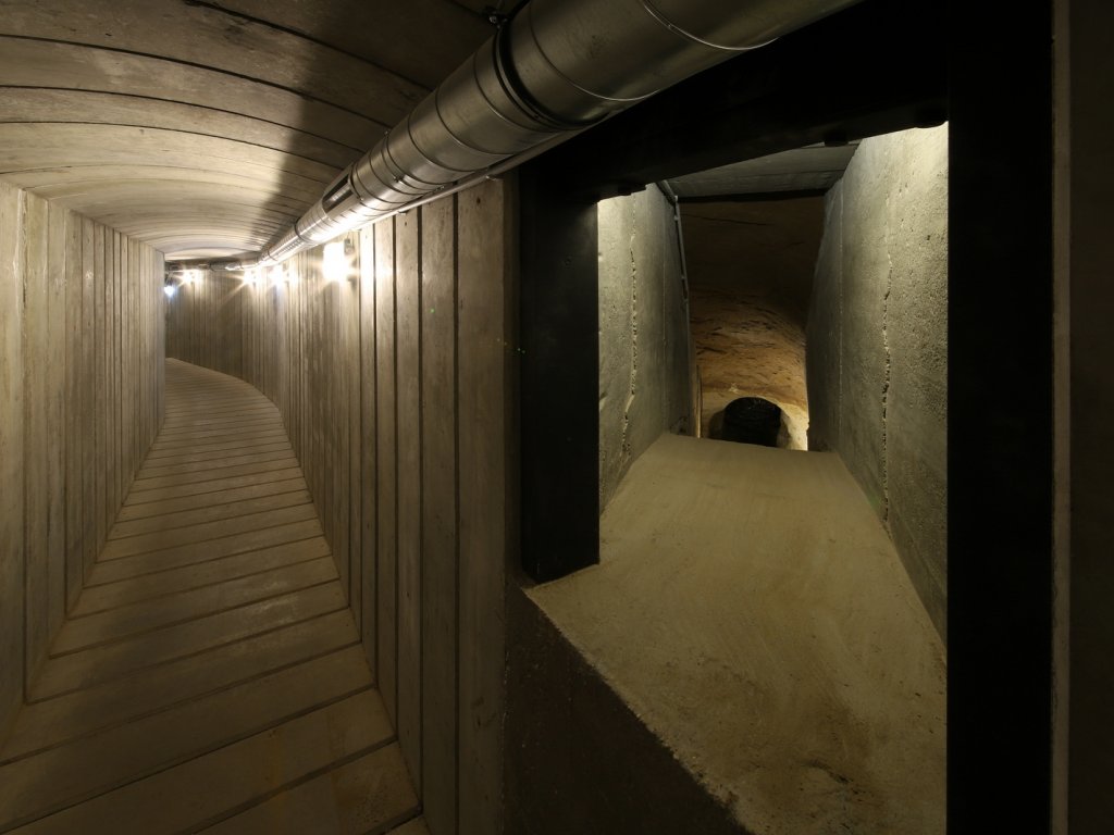 Links befindet sich ein hell ausgeleuchteter, rechteckig verlaufender Betontunnel, der in einer Linksbiegung verschwindet. Davon nach rechts abgehend ist ein Schacht zu erkennen, der den Blick auf einen bogenförmigen, handgegrabenen Tunnel im Bildhintergrund freigibt.