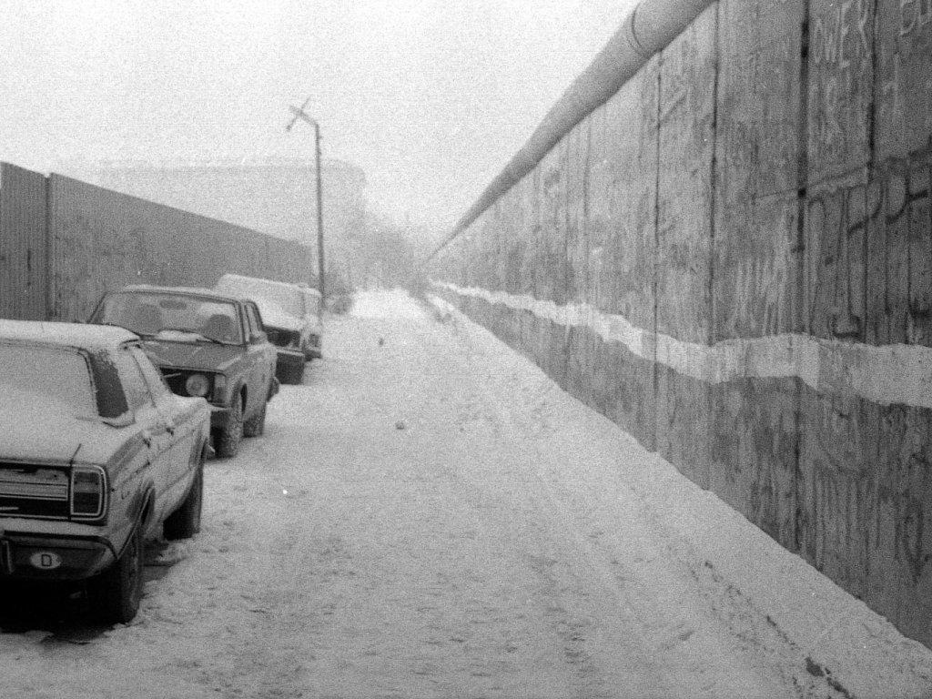 Das Bild entlang der Mauerflucht aufgenommen. Links stehen parkende Autos. Rechts die Berliner Mauer, in deren unterem Drittel ein weißer Strich verläuft. Der Gehweg ist verschneit, der Himmel neblig.