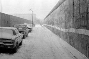Das Bild entlang der Mauerflucht aufgenommen. Links stehen parkende Autos. Rechts die Berliner Mauer, in deren unterem Drittel ein wei&szlig;er Strich verl&auml;uft. Der Gehweg ist verschneit, der Himmel neblig.