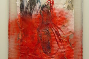 Nahaufnahme eines Gem&auml;ldes. Das Bild besteht aus einer auf Rohrrahmen aufgespannten Plastikfolie. Wei&szlig;es Papier ist darauf angebracht, das teilweise angebrannt und mit Farbe bemalt ist. In roter Farbe ist im unteren Bildteil der Abdruck der Streckmetallzaunes zu erkennen.