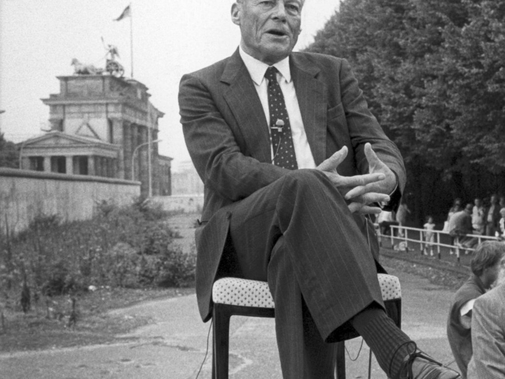 1986: Willy Brandt wird am 25. Jahrestag des Mauerbaus vor der Mauer am Brandenburger Tor von einem amerikanischen Fernsehsender interviewt. Rechts davon befindet sich das Reichstagsgebäude, in dem eine offizielle Gedenkveranstaltung stattfindet.
