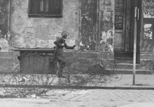 Auf der Straße vor einer heruntergekommenen Häuserfassade verläuft ein Stacheldrahtverhau. Schumann mit Helm und Gewehr setzt von links kommend zum Sprung über die Absperrung an.