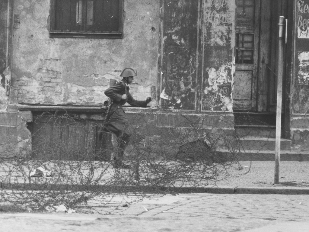 Auf der Straße vor einer heruntergekommenen Häuserfassade verläuft ein Stacheldrahtverhau. Schumann mit Helm und Gewehr setzt von links kommend zum Sprung über die Absperrung an.