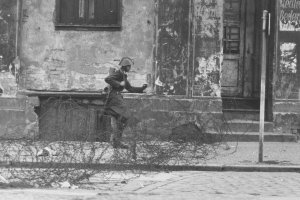 Auf der Straße vor einer heruntergekommenen Häuserfassade verläuft ein Stacheldrahtverhau. Schumann mit Helm und Gewehr setzt von links kommend zum Sprung über die Absperrung an.