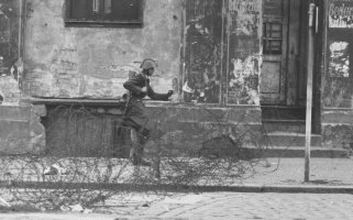 Auf der Straße vor einer heruntergekommenen Häuserfassade verläuft ein Stacheldrahtverhau. Schumann mit Helm und Gewehr setzt von links kommend zum Sprung über die Absperrung an.