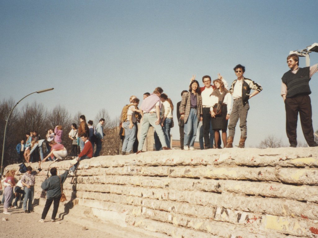 Zahlreiche junge Menschen stehen auf und neben der Panzermauer. Links im Hintergrund ist ein Teil des Reichstagsgebäudes zu erkennen. Die Sonne scheint und die Stimmung wirkt heiter.