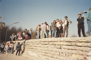 Zahlreiche junge Menschen stehen auf und neben der Panzermauer. Links im Hintergrund ist ein Teil des Reichstagsgebäudes zu erkennen. Die Sonne scheint und die Stimmung wirkt heiter.