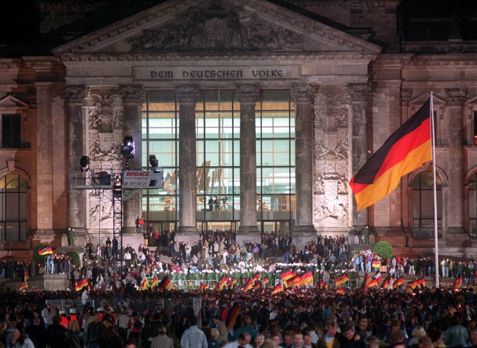 Der Reichstag ist hell beleuchtet, eine Menschenmenge steht vor dem Gebäude, viele schwenken Deutschlandfahnen. Eine große Deutschlandflagge weht rechts im Bild.