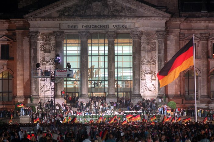 Der Reichstag ist hell beleuchtet, eine Menschenmenge steht vor dem Gebäude, viele schwenken Deutschlandfahnen. Eine große Deutschlandflagge weht rechts im Bild.