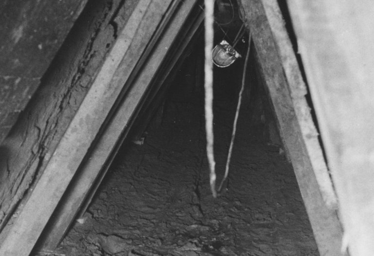 Das Bild ist frontal in die dreieckige Tunnelöffnung aufgenommen. Der Boden ist schlammig, an den Stützbalken hängt eine Lampe.