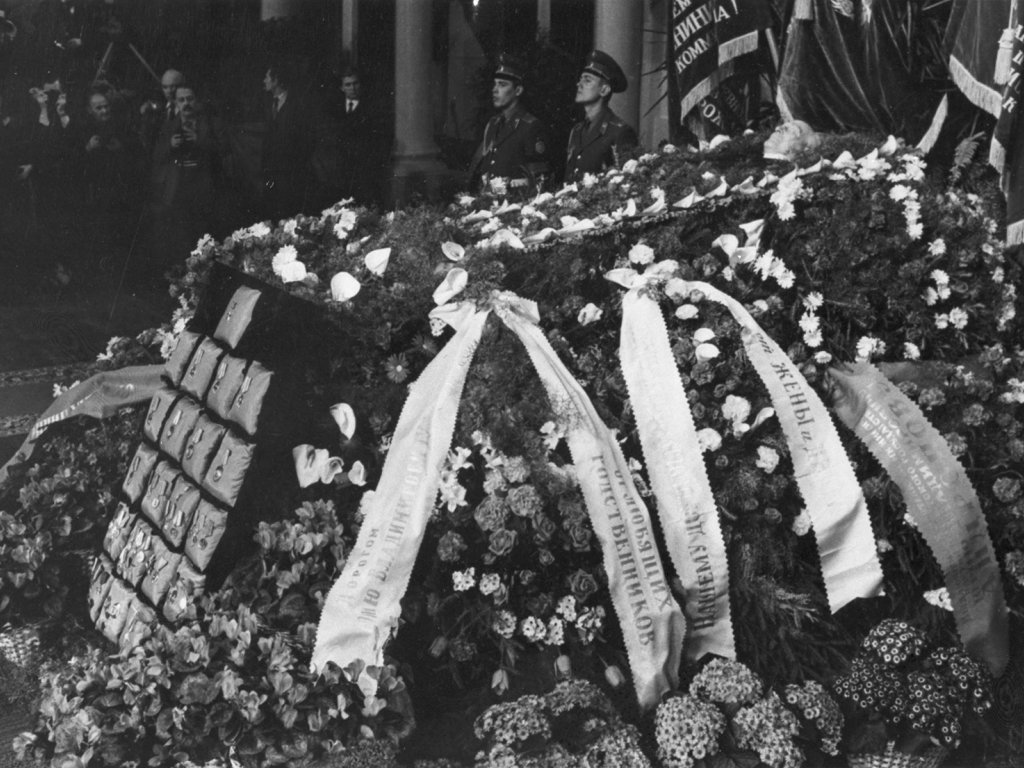 Der Sarg Juri Andropows im Säulensaal des Hauses der Gewerkschaften, Moskau, 12. Februar 1984 Aufnahme vom offenen Sarg, der mit Orden und Blumenkränzen mit Gedenkbändern bedeckt ist. Im Hintergrund stehen Soldaten und Zivilisten, teilweise mit Kameras.