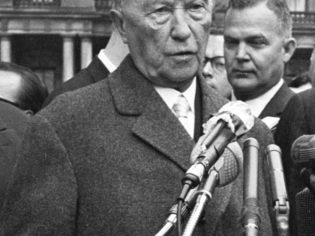 Konrad Adenauer steht mit ernstem Blick vor Mikrofonen und spricht.
