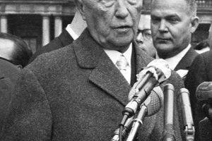 Konrad Adenauer steht mit ernstem Blick vor Mikrofonen und spricht.