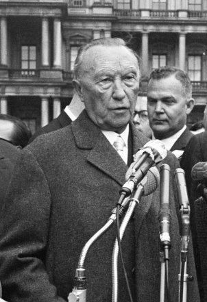 Konrad Adenauer steht mit ernstem Blick vor Mikrofonen und spricht.