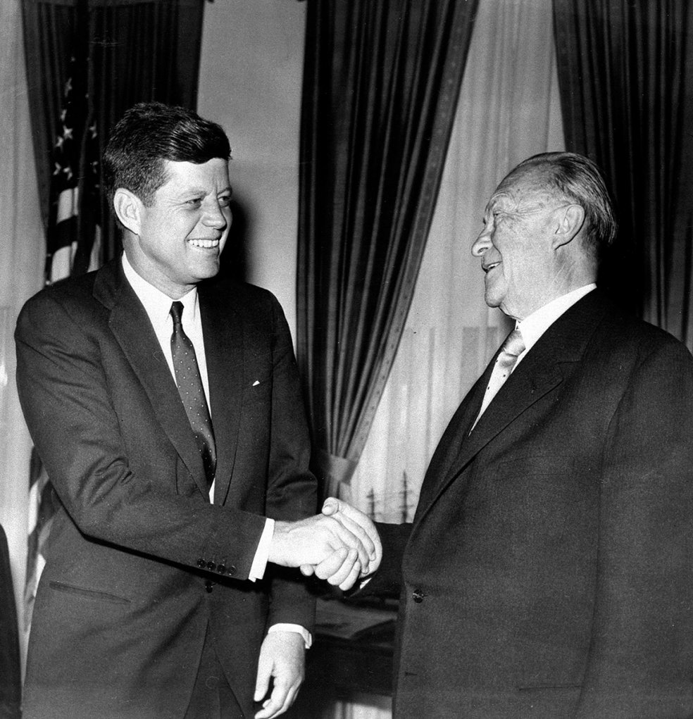 Kennedy steht links im Bild vor einer amerikanischen Flagge, l&auml;chelt und sch&uuml;ttelt dem ebenfalls l&auml;chelnden Adenauer die Hand.