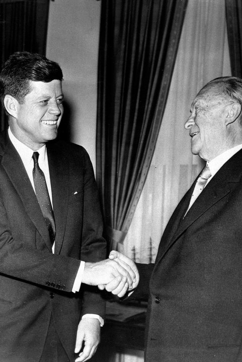 Kennedy steht links im Bild vor einer amerikanischen Flagge, lächelt und schüttelt dem ebenfalls lächelnden Adenauer die Hand.