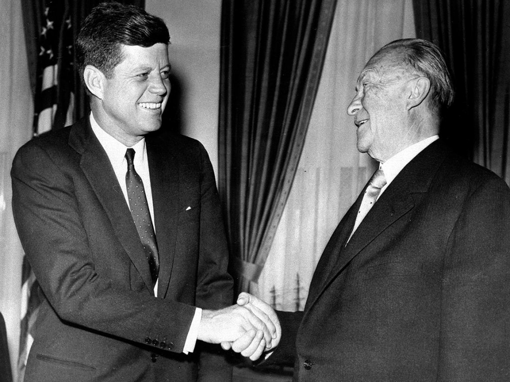 Kennedy steht links im Bild vor einer amerikanischen Flagge, lächelt und schüttelt dem ebenfalls lächelnden Adenauer die Hand.