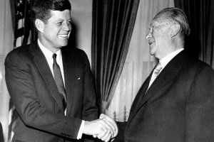 Kennedy steht links im Bild vor einer amerikanischen Flagge, lächelt und schüttelt dem ebenfalls lächelnden Adenauer die Hand.