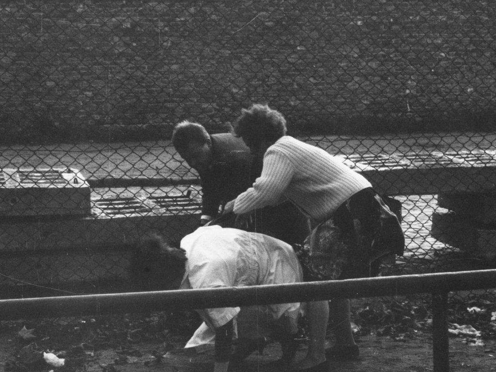 Eine Ost-Berlinerin flüchtet mit Hilfe von Verwandten in den Westen, Berlin-Neukölln, 22. September 1961. Drei Menschen bücken sich vor einer Absperrung aus Betonplatten und Maschendrahtzaun. Im Hintergrund sind eine Mauer und Wohnhäuser zu erkennen.