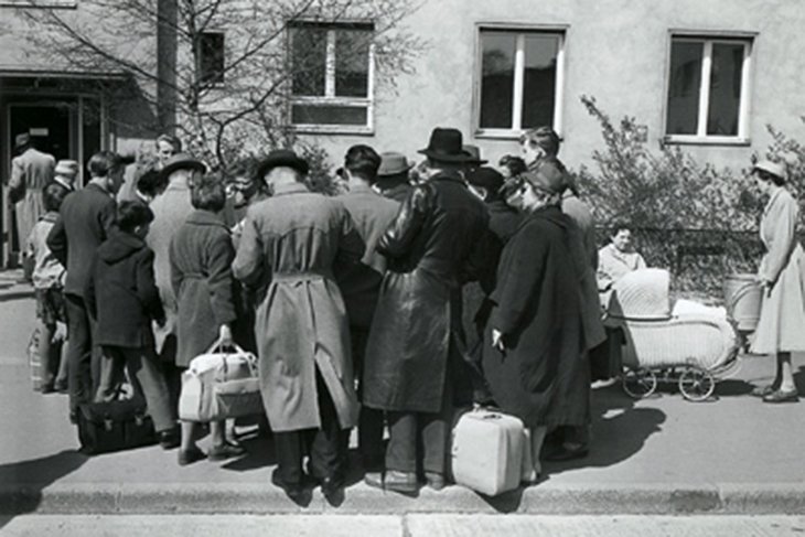 Eine Menschenansammlung steht vor einem Gebäude. Die Rücken sind der Kamera zugewandt, einige haben Gepäck dabei. Rechts ist ein Kinderwagen zu sehen.