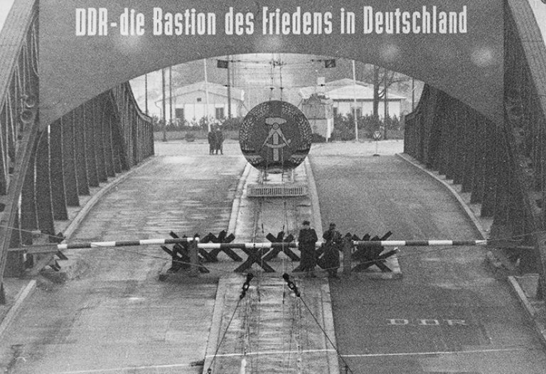 Vogelperspektive auf die Bösebrücke, die mit Panzersperren und Schlagbäumen gesperrt ist. Mittig stehen Soldaten. Die Brücke ist beschriftet mit: DDR – die Bastion des Friedens in Deutschland. Unter dem Schriftzug hängt das DDR-Staatswappen.