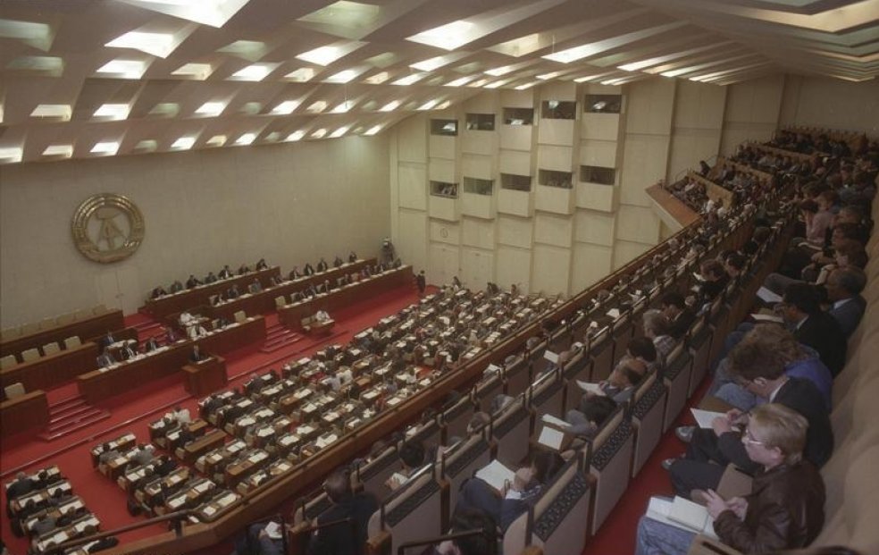 Der Plenarsaal der Volkskammer von der hinteren linken Ecke der Empore aus aufgenommen. Der fensterlose mit rotem Teppich ausgelegte Saal ist voll, fast alle Sitze sind besetzt. An der Wand prangt das goldene Emblem der DDR.