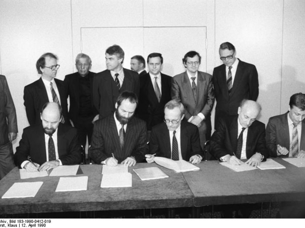 Die an der neuen DDR-Regierung beteiligten Parteien unterzeichnen im Hause der Volkskammer die Koalitionsvereinbarung zwischen den Fraktionen der CDU, der DSU, dem DA, den Liberalen (FDP, BFD, F.D.P.) und der SPD vom (v. l. n. r.: Rainer Eppelmann, Markus Meckel, Lothar de Maiziere, Hans-Wilhelm Ebeling und Prof. Dr. Rainer Ortleb); Aufnahme 12. April 1990  An einem langen Tisch sitzen die fünf Fraktionsvertreter und unterzeichnen die vor ihnen liegenden Papiere. Hinter ihnen stehen acht Männer und beobachten das Geschehen.